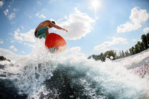 Young sports guy jumping on bright wakeboard down the river water against the background of sky