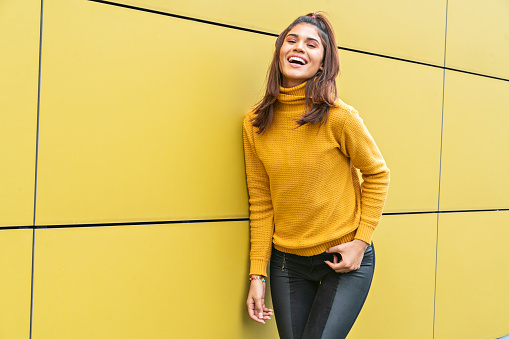 Beautiful Latina woman between 20 and 25 years old looks bored on the street with yellow clothes leaning against a yellow wall during the COVID-generated quarantine 19