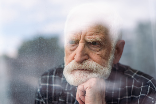 grieving senior man looking away through window glass while holding hand near chin