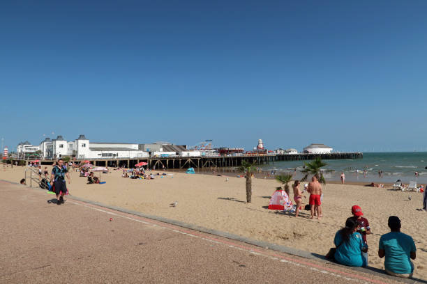 Beach with pier in the background at Clacton in Essex Beach and promenade at Clacton with Victorian pleasure pier in the background. Holiday makers sunbathing on the beach. Outdoors on a sunny summers day. Clacton on Sea, Essex, United Kingdom, July 17, 2020 clacton on sea stock pictures, royalty-free photos & images