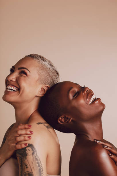 Beautiful women standing together and smiling Happy diverse female models standing back to back with their head touching. Two women standing together and smiling over beige background. shaved black women stock pictures, royalty-free photos & images