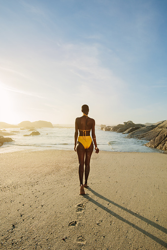 Rear view of a african woman wearing yellow bikini walking into the sea. Woman walking towards sea in swimwear on a summer day.