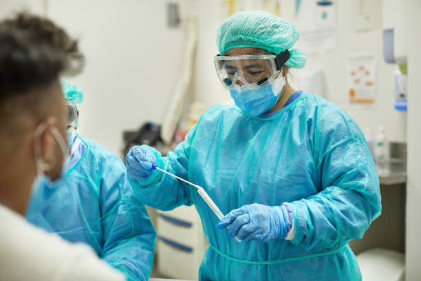 Hospital Nurse Placing Test Swab in Transport Medium Over the shoulder view of healthcare worker in protective clothing, eyewear, bouffant cap, surgical mask, and gloves placing swab specimen in sterile container. frontline worker mask stock pictures, royalty-free photos & images