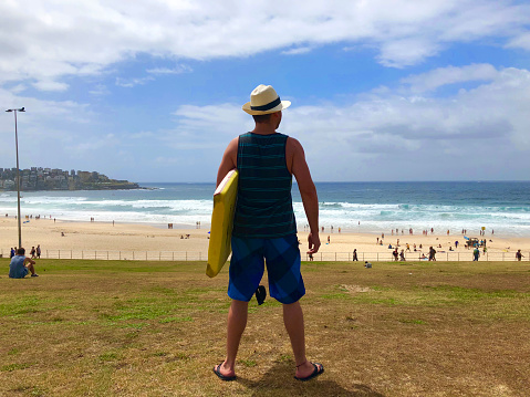 Young handsome Latin male tourist going to surf carrying his board in the most famous beach around Australia: Bondi Beach in  Sydney, New South Wales, wearing a panama hat, tank top, board shorts and sandals.