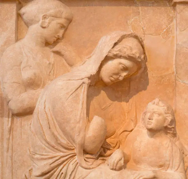 Photo of Ancient bas-relief depicting young seated woman with boy and maiden stands in background on ancient Greek funerary stele from Kerameikos, Athens, Greece