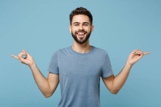istock Young handsome smiling funny man in casual clothes posing isolated on blue background, studio portrait. People sincere emotions lifestyle concept. Mock up copy space. Pointing index fingers aside. 1256899234