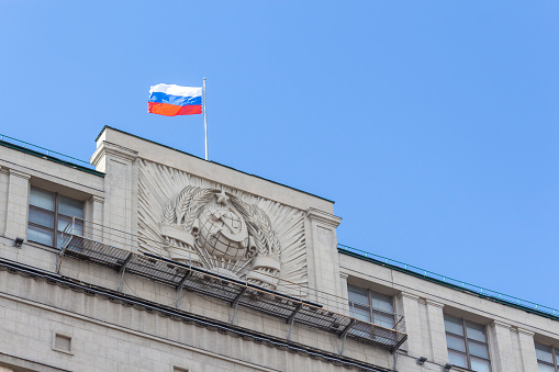 Russian National flag on top of the State Duma building, also known as Gosduma. Soviet coat of arms on the wall of building. Clear blue sky.