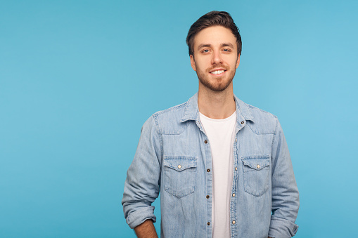 Portrait of happy handsome young man with stylish hairdo and bristle, wearing worker denim shirt looking at camera with attractive smile, positive outlook. studio shot isolated on blue background