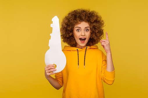 Portrait of inspired, amazed curly-haired woman in urban style hoodie pointing finger up with genius idea and holding big paper key, looking surprised by sudden solution. indoor studio shot isolated