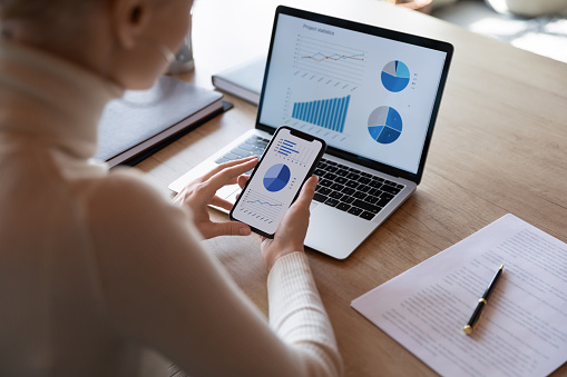 Woman hold smartphone use pc at workplace. Project stats financial data sales charts on laptop and cellphone screen, close up view over shoulder. Report preparation, synchronization for safety concept