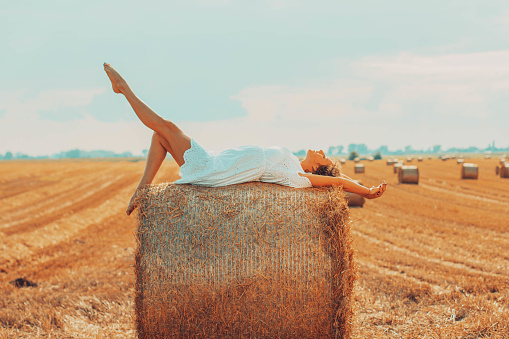 Young woman on a hay bale while overlooking the sky during harvest season.
