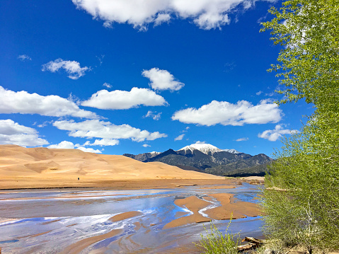 The snow capped Sangre de Cristo Mountains rise in the distance beyond the sand dunes of the Great Sand Dunes National Park and Preserve and Medano Creek in southern Colorado on a beautiful cloud filled day.