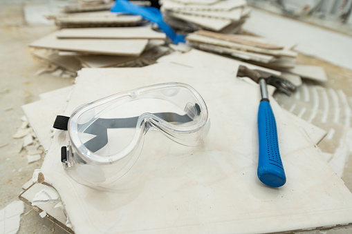 This still life photograph is a close up of clear safety goggles and the hammer tool used to remove the stack of tile flooring in an apartment kitchen during home renovation.