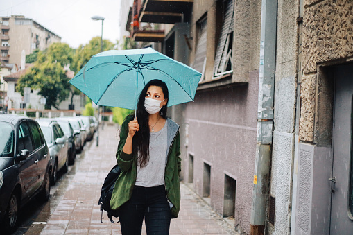 Young woman getting caught in the rain while walking in the city centre, wearing a protective face mask during the coronavirus pandemic.