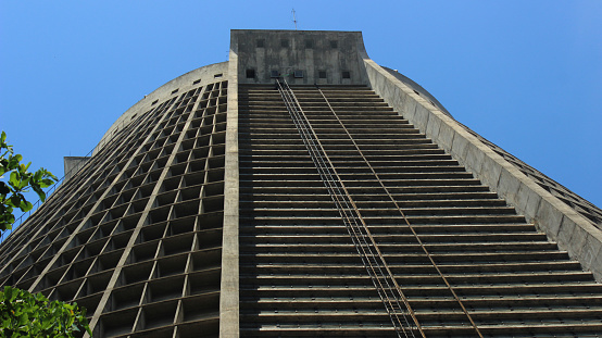 Rio de Janeiro, Brazil: Low angle view of the Metropolitan Cathedral (brutalist architecture), designed as a pyramid with reinforced concrete, some trees on the sides and a blue sky of a hot summer day