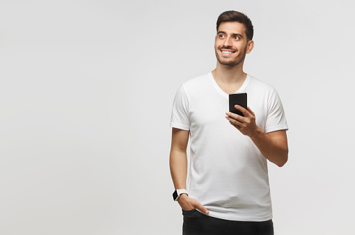 Banner of young man standing in white t-shirt isolated on gray background, holding smartphone in hand and looking aside with dreamful smile