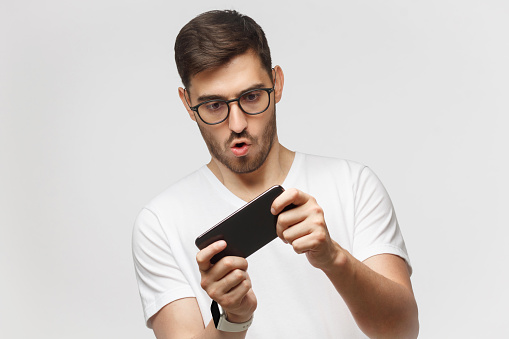 Young man in white t-shirt holding phone with both hands, moving it while playing video game in app, making funny face, isolated on gray background