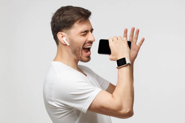 Young handsome man singing his favorite song, using his phone instead microphone, wearing white t-shirt and wireless earphones