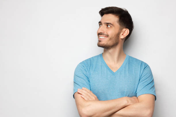 istock Young handsome man in blue tshirt, standing with crossed arms, feeling confident, looking away to copy space, isolated on gray background 1256944008