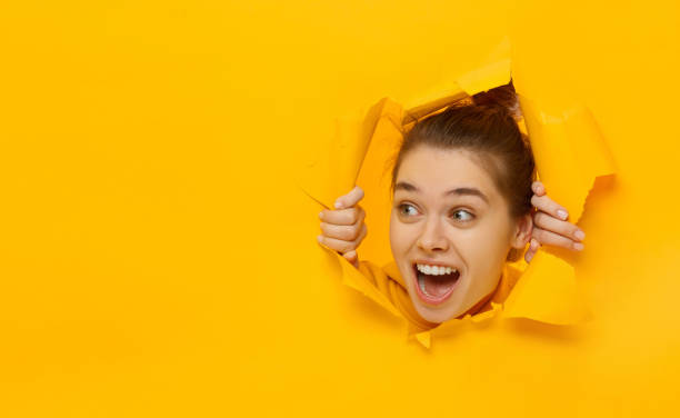 Excited girl looking through hole in paper at left with eyes round with surprise, isolated on yellow background with copy space Excited girl looking through hole in paper at left with eyes round with surprise, isolated on yellow background with copy space peeking photos stock pictures, royalty-free photos & images