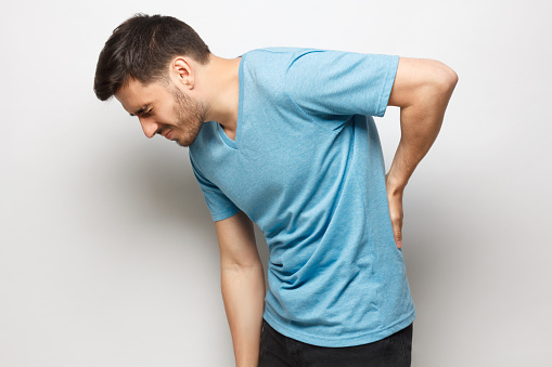 Young unhappy guy in blue t-shirt suffering from severe backache, touching back trying to ease pain in spine, isolated on gray background