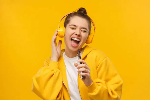 Photo of Trendy girl singing favorite song out loud in phone as mic, wearing wireless headphones, isolated on yellow background. Karaoke online app.