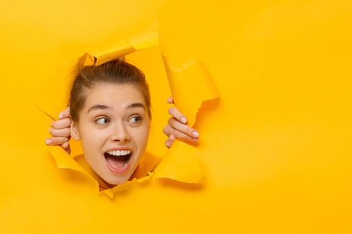 Young girl tearing paper and peeking out hole, curious about something on copy space on right, isolated on yellow background