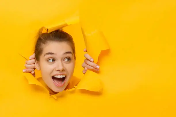 Photo of Young girl tearing paper and peeking out hole, curious about something on copy space on right, isolated on yellow background