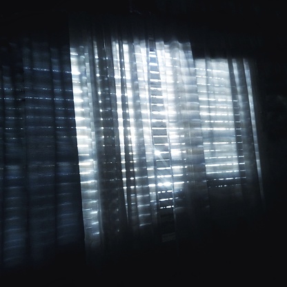 Morning sunlight coming through some old wooden shutters and dark curtains in a dark room during quarantine. A photo that shows sadness, loneliness but hope during a night of insomnia.