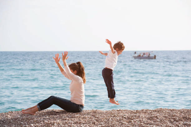 healthy family sitting mother and jumping high kid on sea shore in autumn sunset - small gymnastics athlete action imagens e fotografias de stock