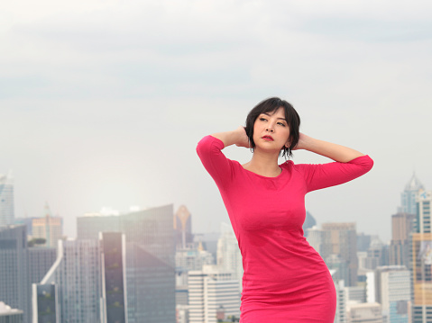 Portrait of cheerful confident young female standing with skyscraper city view. Attractive woman in red dress on rooftop