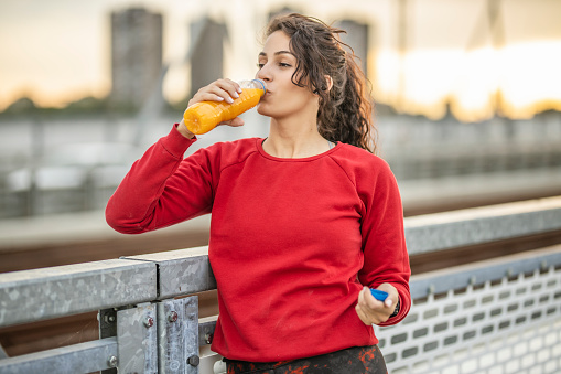 Thirsty sporty woman resting and drinking orange juice after jogging on bridge