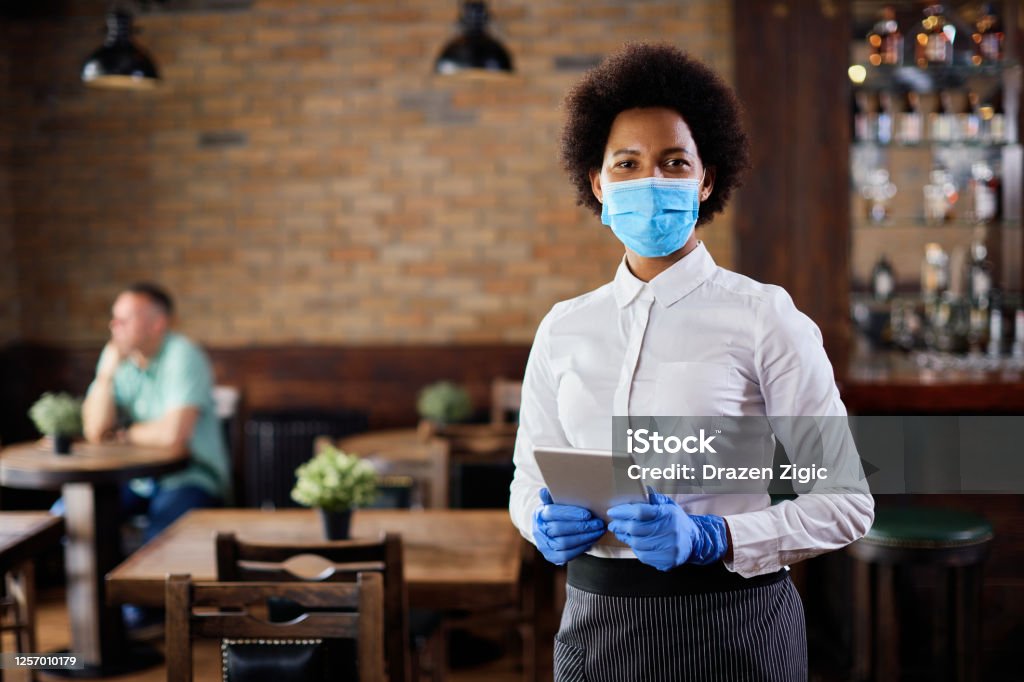 Portrait of African American waitress with protective mask in a cafe. Portrait of black waitress wearing protective face mask while holding touchpad and looking at the camera in a pub. Restaurant Stock Photo