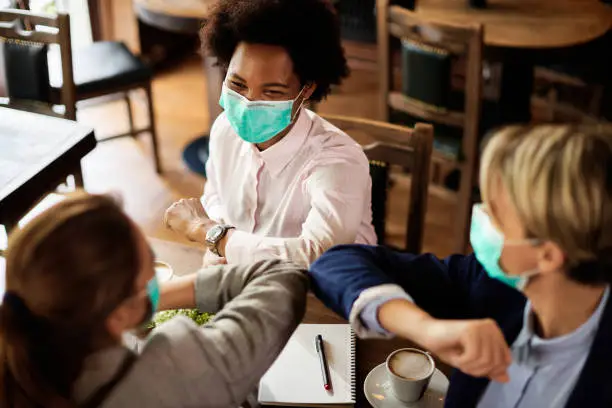 Group of happy female colleagues with protective face masks greeting with elbows in a cafe.