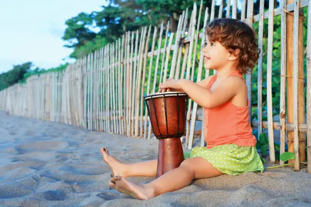 Little happy baby girl play ethnic music on traditional african hand drum djembe, enjoying sunset on ocean beach. Children healthy lifestyle. Travel, family activity on tropical island summer holiday.