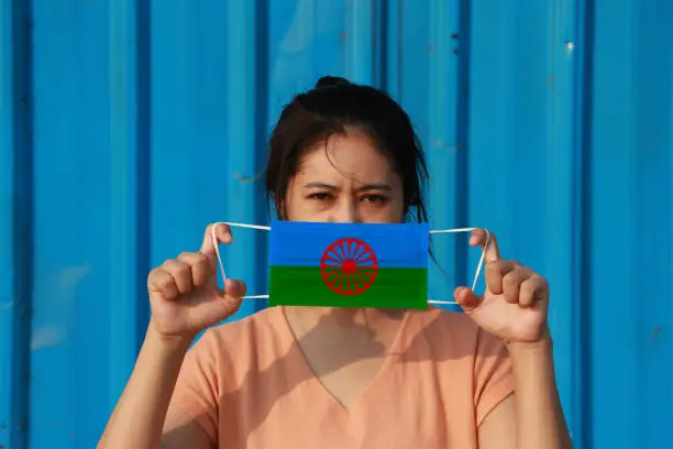 A woman with Romani people flag on hygienic mask in her hand and lifted up the front face on blue background. Tiny Particle or virus corona or Covid 19 protection. Concept of Combating illness.