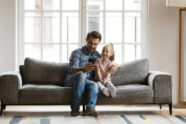 istock Happy father with little daughter using smartphone, sitting on couch 1257033419