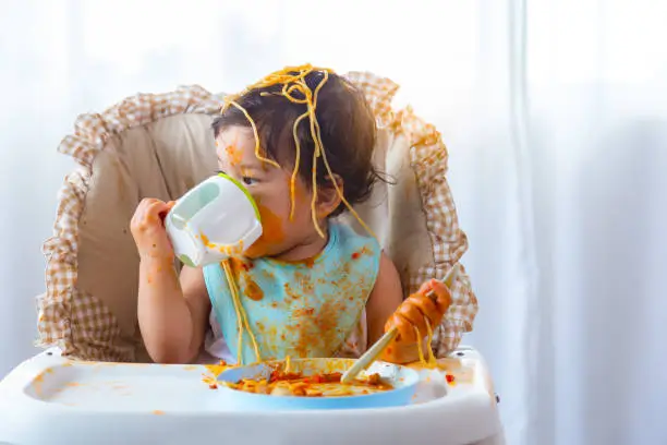 Photo of Adorable little toddler girl or infant baby drink water after eat delicious spaghetti food on chair. Funny cute infant girl get thirsty. Lovely mix race daughter get dirty. Kid hold spoon upside down