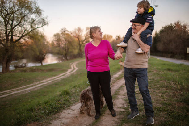 Grandparents And Grandson Walking with dog Grandfather Giving Grandson Ride On Shoulders As They Walk With Grandmother and their dog by the river. dog disruptagingcollection stock pictures, royalty-free photos & images