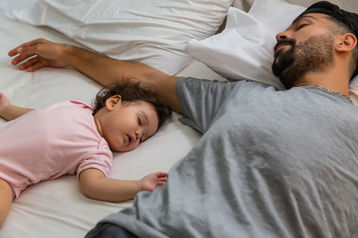 Little girl sleeping on bed near her father in bedtime. Single father get tired and sleeping together with his beloved infant daughter. Adorable little baby girl had good sleep and get comfortable