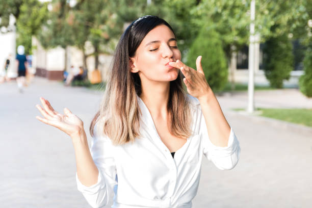 portrait of beautiful woman in white shirt and hair bezel with mouth full of food licking her fingers outdoor in city park and enjoying junk but tasty fast food while walking portrait of beautiful woman in white shirt and hair bezel with mouth full of food licking her fingers outdoor in city park and enjoying junk but tasty fast food while walking. chocolate fingers stock pictures, royalty-free photos & images