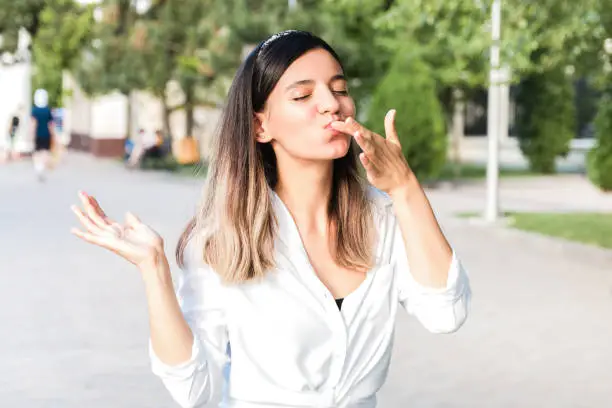 portrait of beautiful woman in white shirt and hair bezel with mouth full of food licking her fingers outdoor in city park and enjoying junk but tasty fast food while walking.