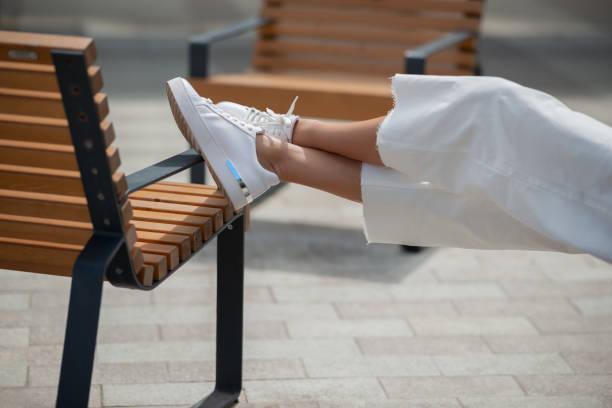 Women's legs in white sneakers and white trousers. Female feet in a city park lying on a bench on a warm summer day. cropped pants photos stock pictures, royalty-free photos & images