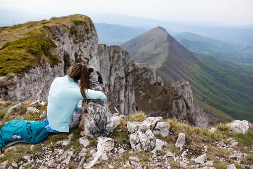 A young woman is sitting on top of a mountain hugging her dog and taking in the view