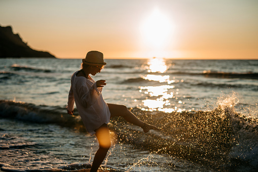 Young playful woman running on beach, splashing sea vawes with leg. Golden hour