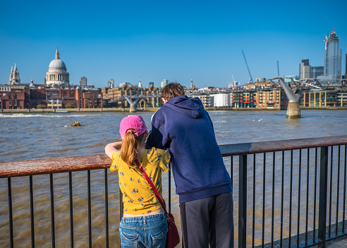 Teenage boy and his preteen sister leaning on parapet of  South Bank of River Thames looking at iconic buildings of city of London, England, on the other side on nice spring day