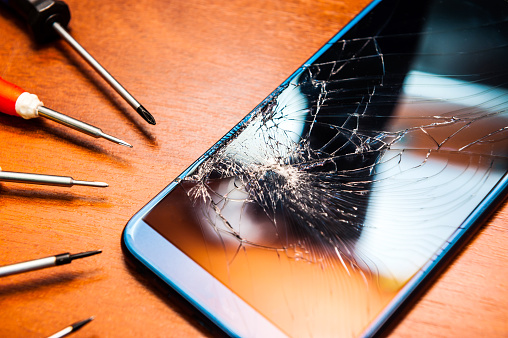 A broken mobile phone with a broken screen with tools, screwdrivers on a wooden table. Electronics repair shops. High quality photo