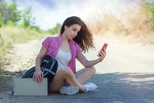 Beautiful young woman hitchhiker with a backpack holds a cardboard with a place for text sitting on the road and looks into the phone. Hitchhiking with a mobile cell phone.
