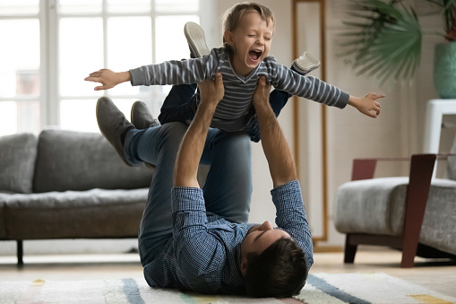 Laughing little boy with hands outstretched pretending flying, playing funny game with happy father, loving young dad holding lifting adorable preschool son, lying on warm floor in living room