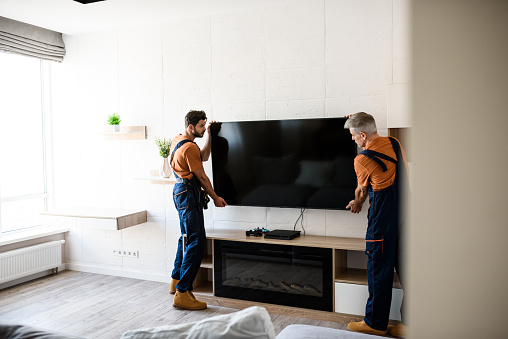 Two handymen, workers in uniform hanging, installing tv television on the wall indoors. Repair and assembly service concept. Selective focus. Horizontal shot